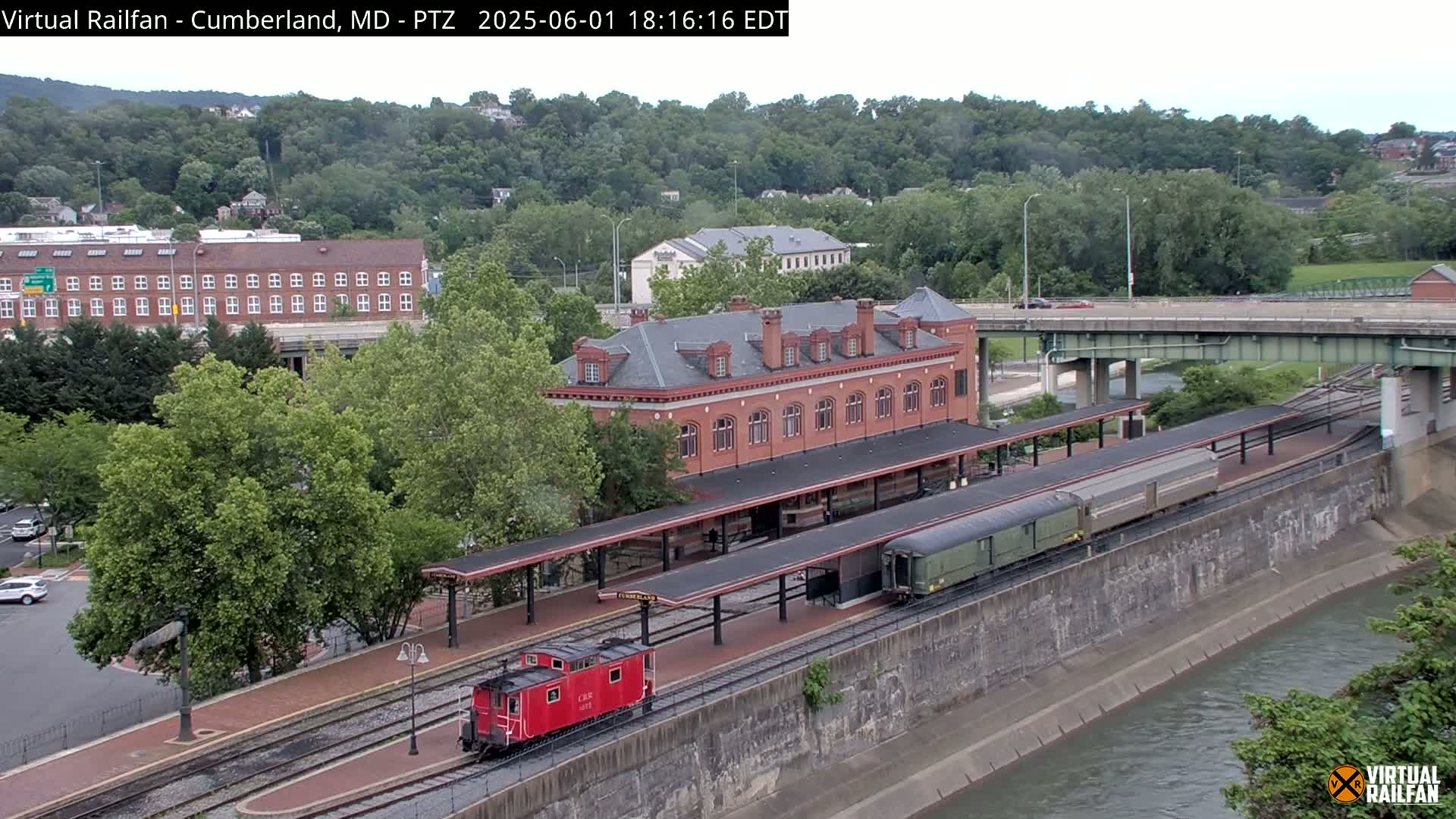 A red train and a long green passenger car sit at a brick train station next to a river, under a partly cloudy sky.