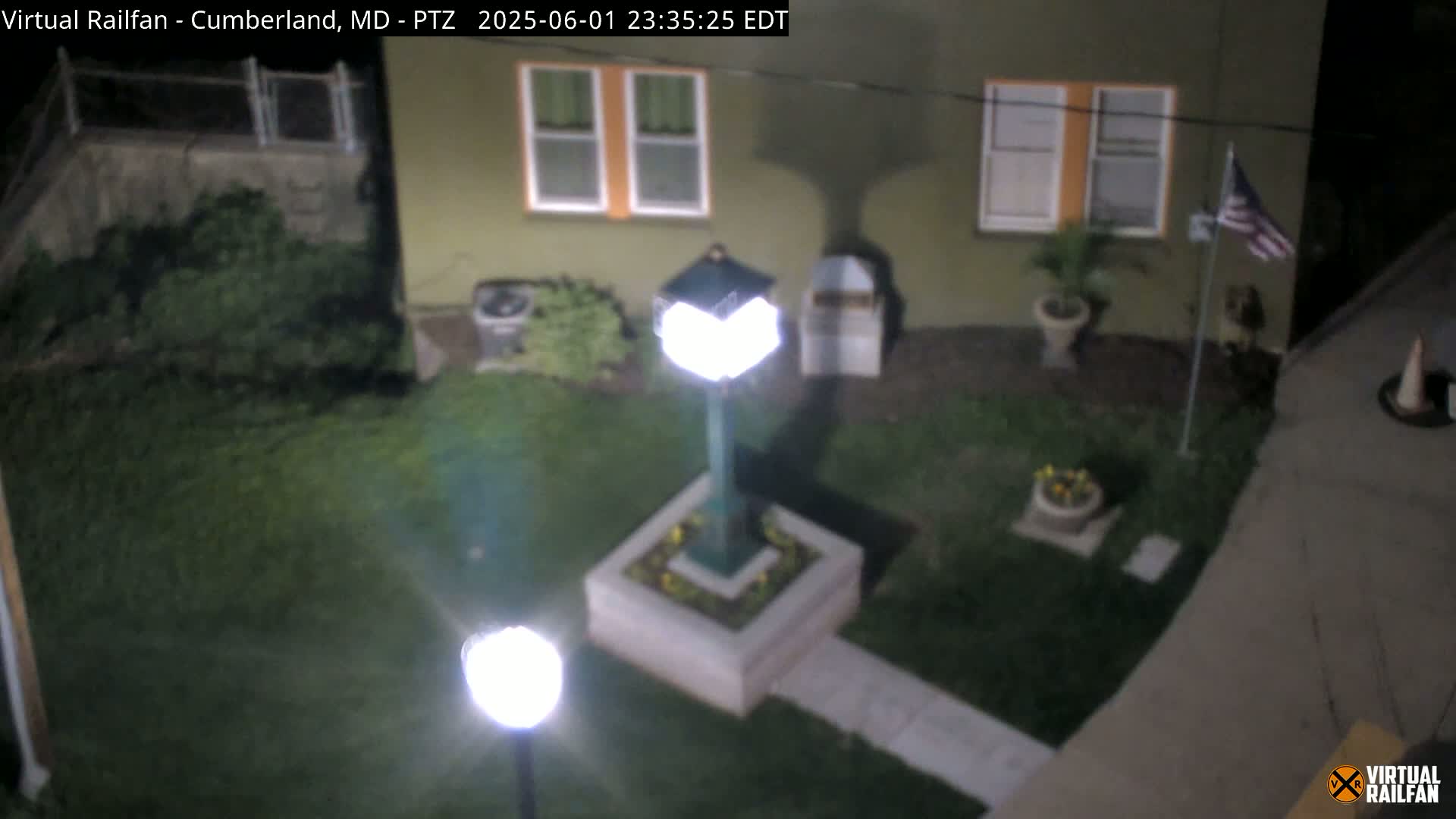 A nighttime, overhead view shows a small, grassy yard with a light post, potted plants, a flagpole, and a section of a building’s exterior.