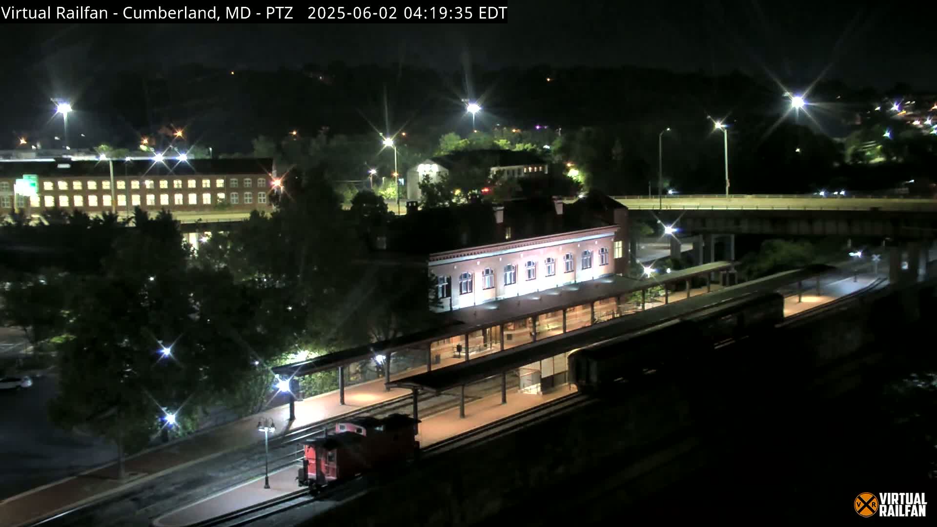 A nighttime view of a train at a station, with a long brick building, and an elevated roadway in the background.