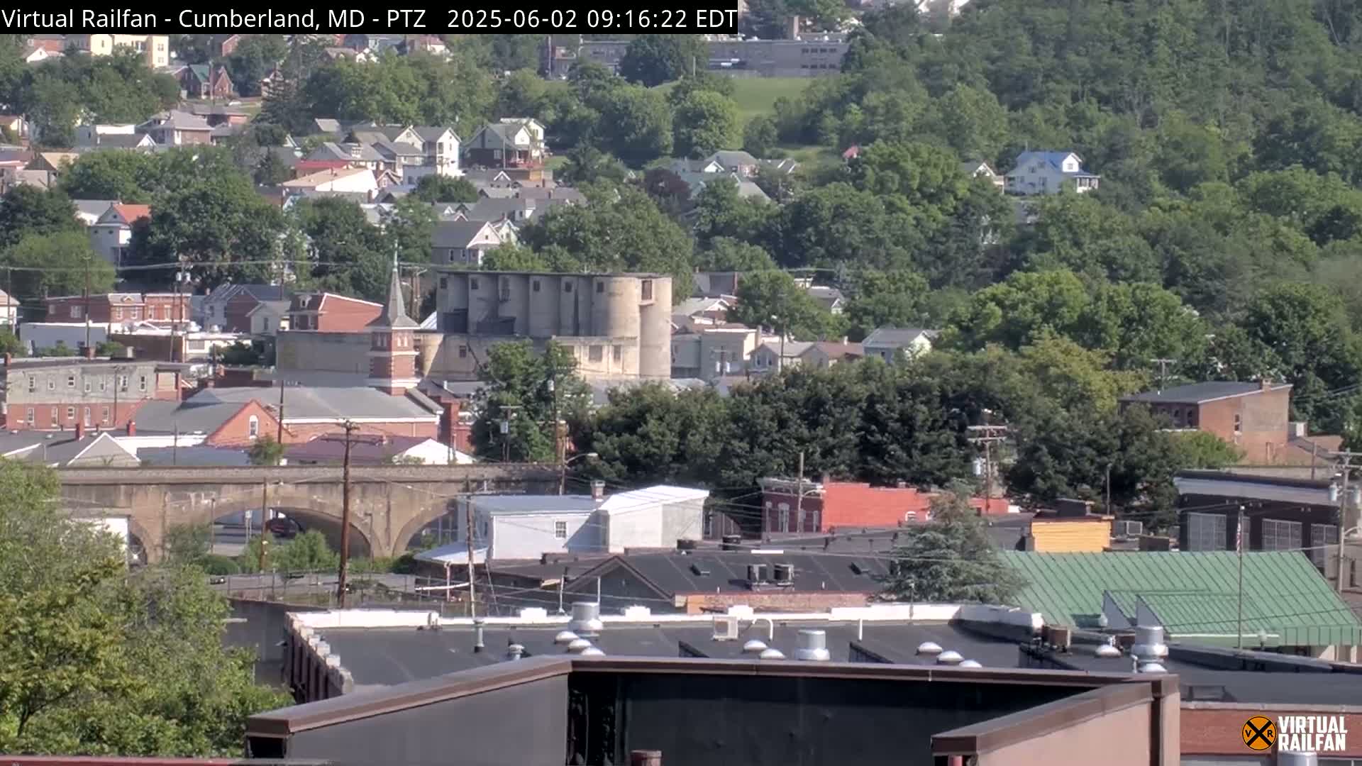 A long shot of a town on a sunny day, featuring a stone bridge, several buildings, and a cluster of grain silos, all surrounded by lush green trees.