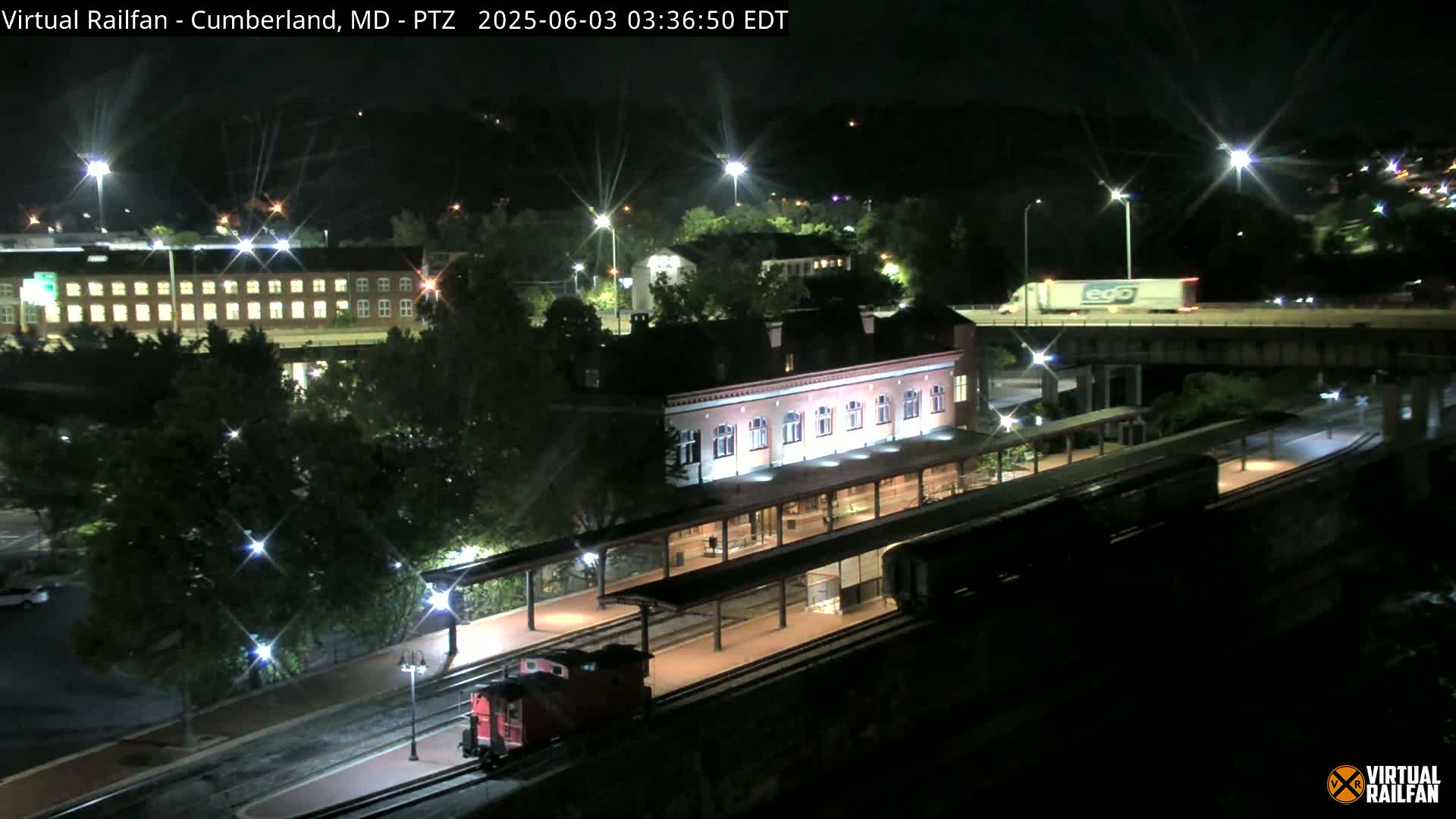 A nighttime view of a train at a station next to a highway overpass, with streetlights illuminating the scene.
