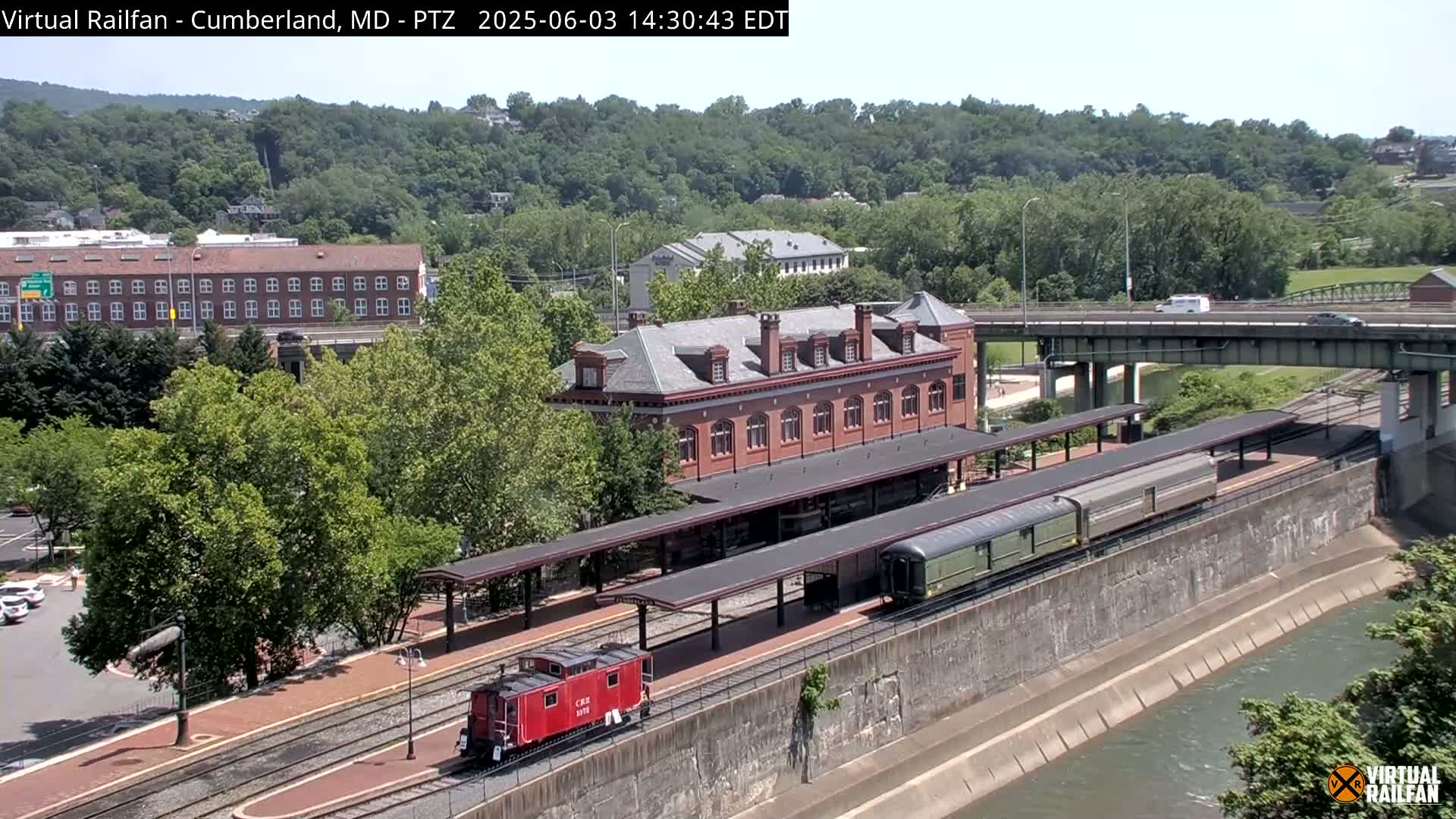 A red caboose and a long passenger train are parked at a brick train station next to a river on a sunny day.