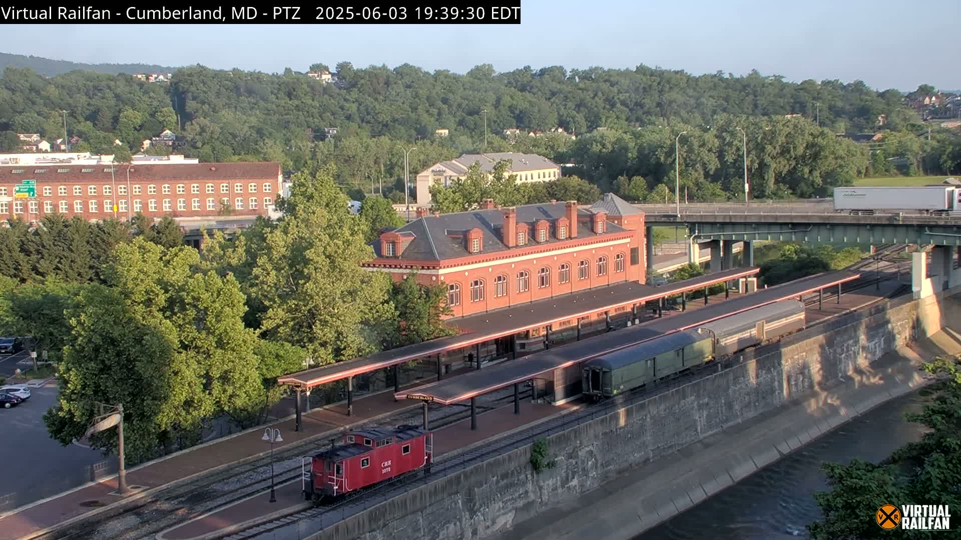 A red caboose and a long train are parked at a brick train station next to a river under a clear, evening sky.