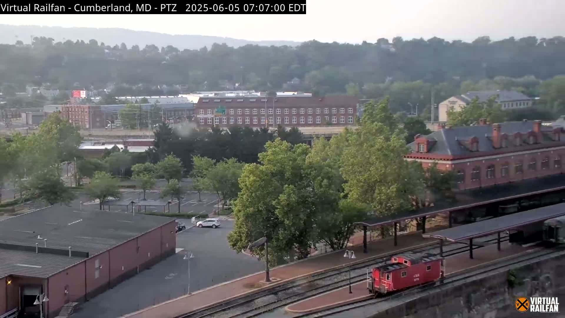 A hazy view of a town with a train station and railway tracks, including a red train car, and a parking lot is visible from an elevated perspective.