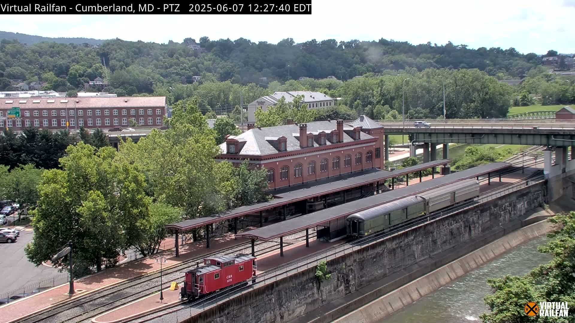 A red caboose and a long passenger train are at a station next to a river, under a partly cloudy sky.