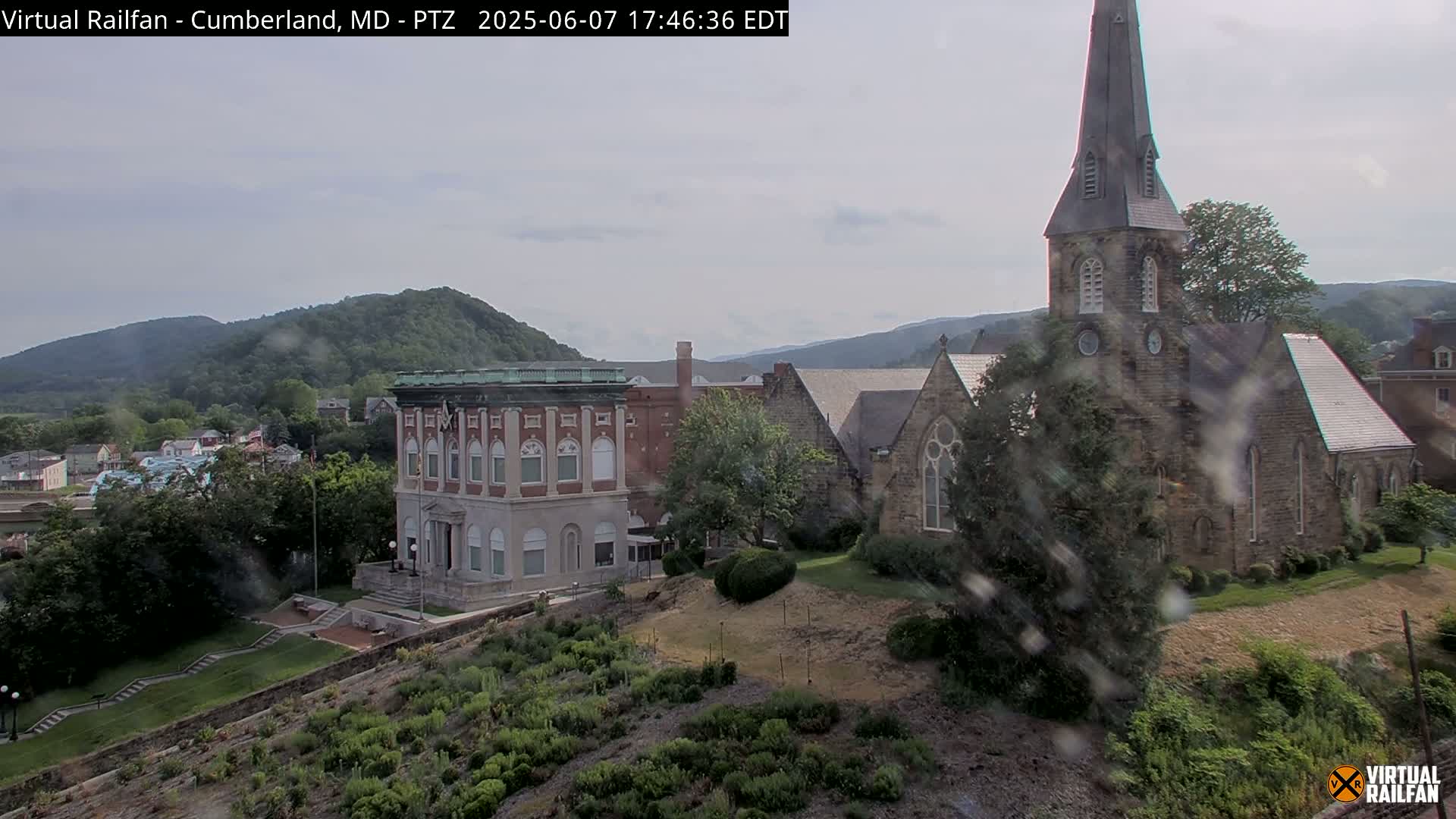 A brick church and a large brick building stand on a hillside overlooking a town nestled in green mountains under an overcast sky.