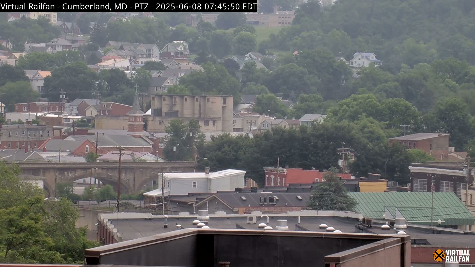 A hazy view of a town situated on a hillside, showing numerous buildings, a stone bridge, and a large grain silo.