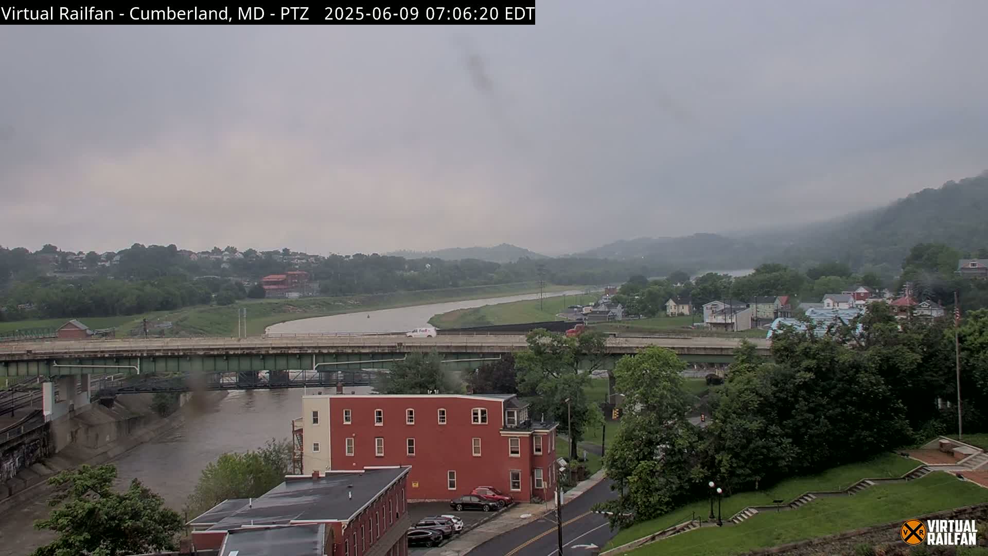 A river flows through a valley under an overcast sky, passing beneath a bridge and alongside buildings and green hills.