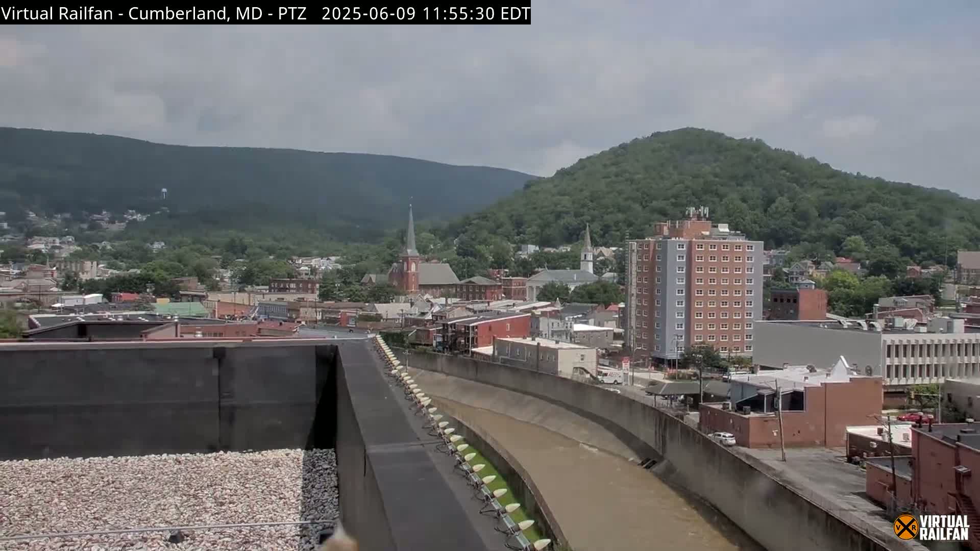 A view of a town nestled in a valley under a partly cloudy sky, with a river curving through the foreground.