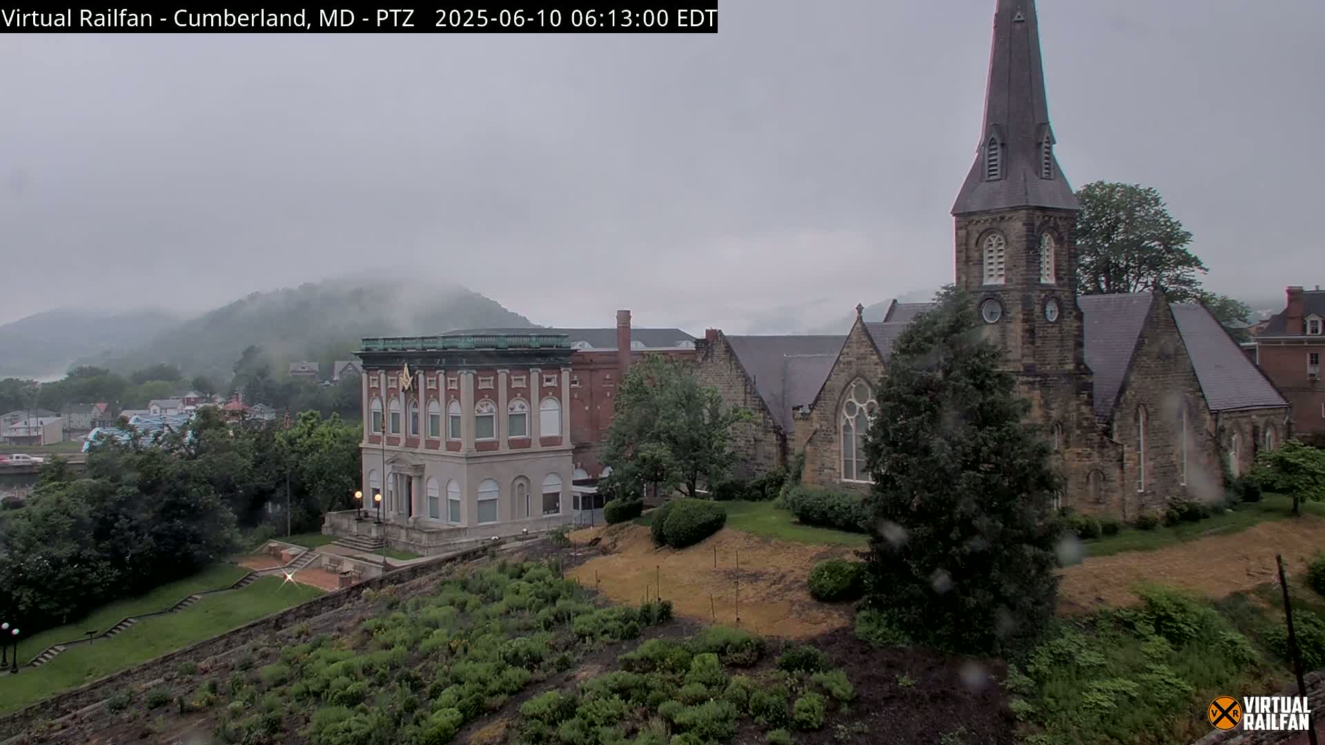 A rain-covered view of a town nestled in a valley, featuring a large brick building and a stone church with a tall steeple, with fog-covered mountains in the background.