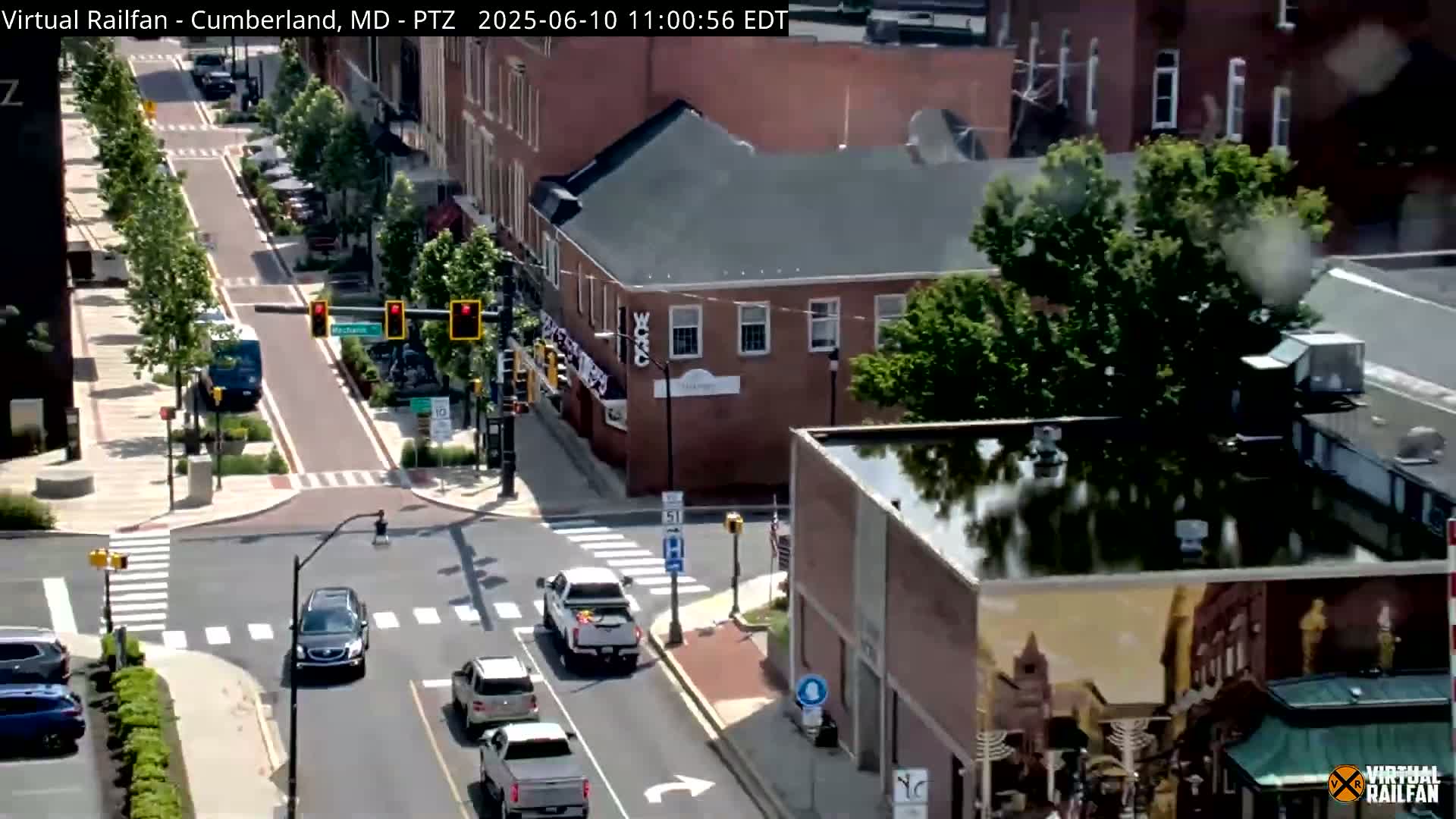 A street scene on a sunny day shows a traffic intersection in a town with brick buildings and green trees, several cars are visible, and a pedestrian crosses the street.