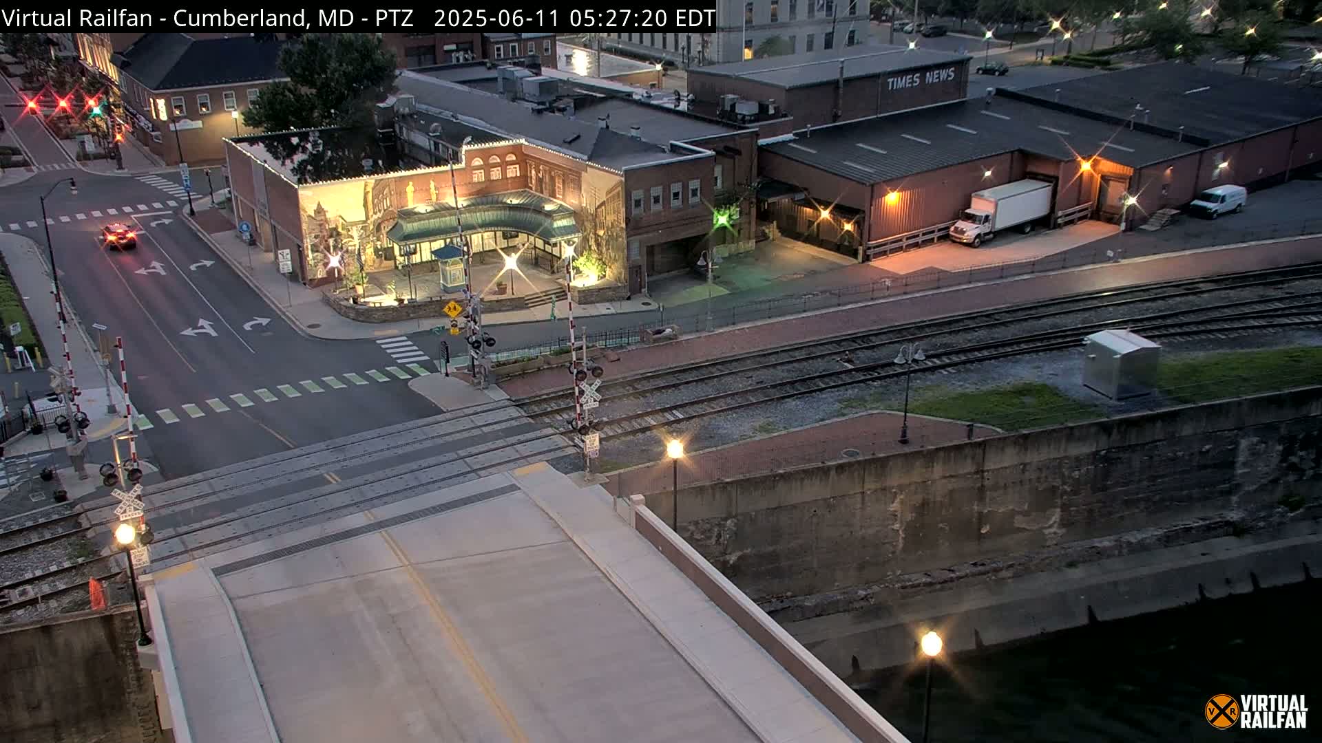 A high-angle, nighttime view of a street intersection with railroad tracks, a bridge, and several buildings, including one with a large mural, under a dark sky.