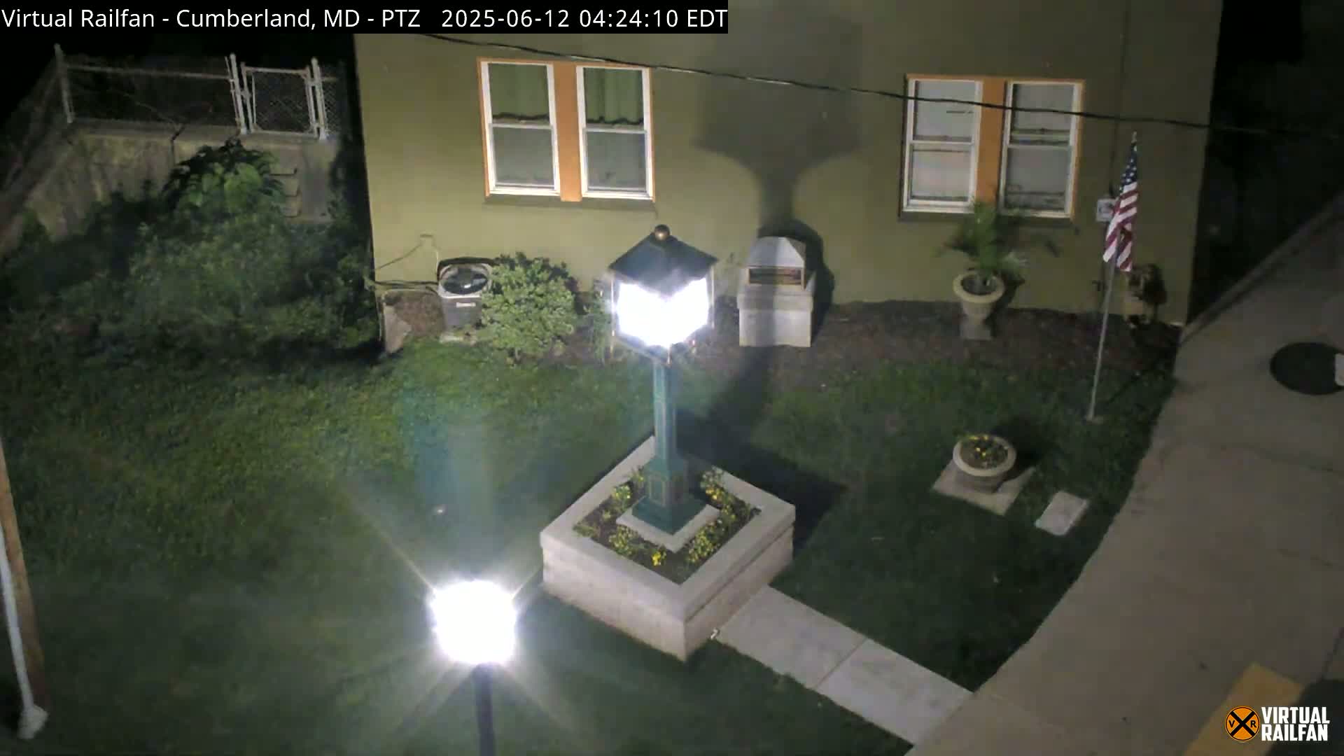 A nighttime, overhead view shows a lit lamp post in a small garden next to a building with an American flag, illuminated by artificial light.
