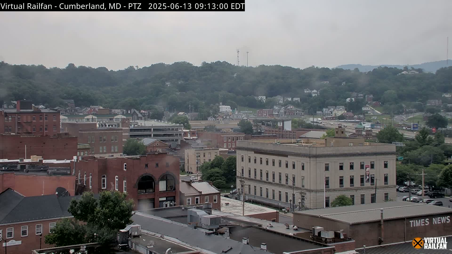 A cloudy day reveals a city nestled in a valley, with brick buildings and a large, light-colored building prominent in the foreground, surrounded by green hills.