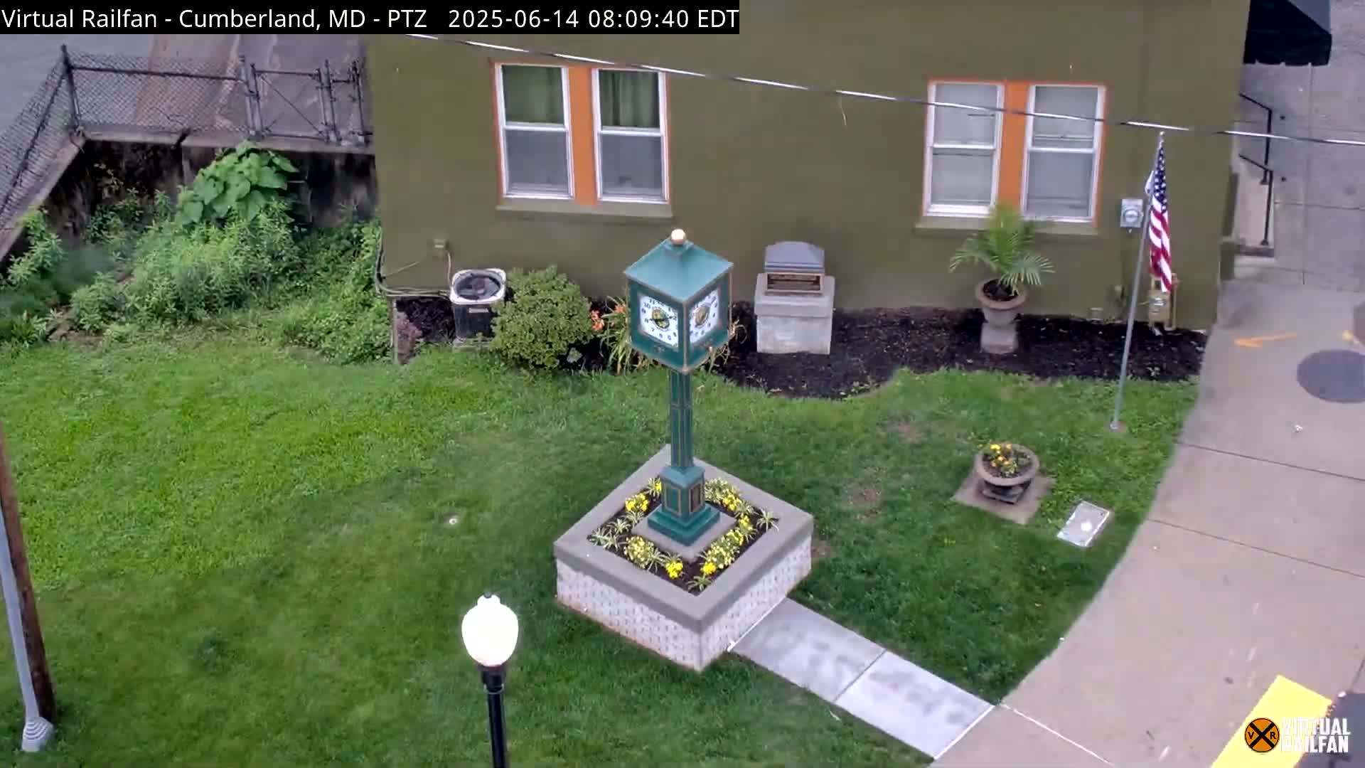 An overhead view shows a green clock tower situated in a small flower bed next to a building, all surrounded by green grass and under a cloudy sky.