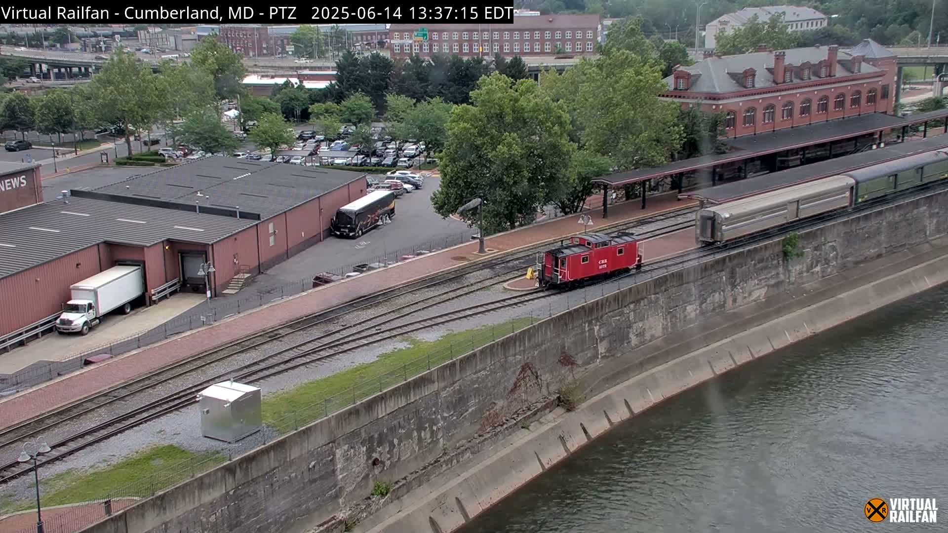 A red caboose and a long passenger train are on railroad tracks next to a river on an overcast day, with buildings and vehicles visible in the background.