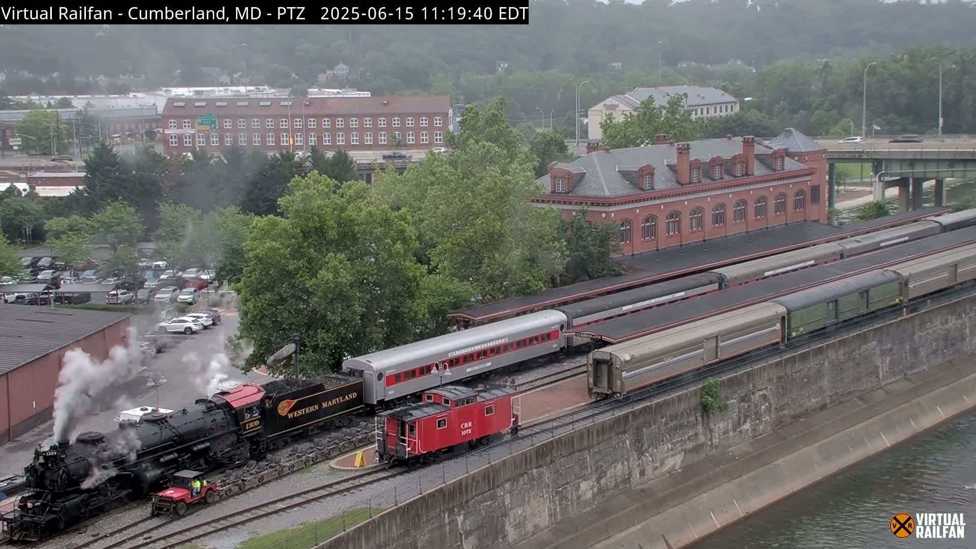 A steam locomotive is emitting smoke near several passenger cars and a red caboose next to a brick building, all situated beside a body of water under an overcast sky.
