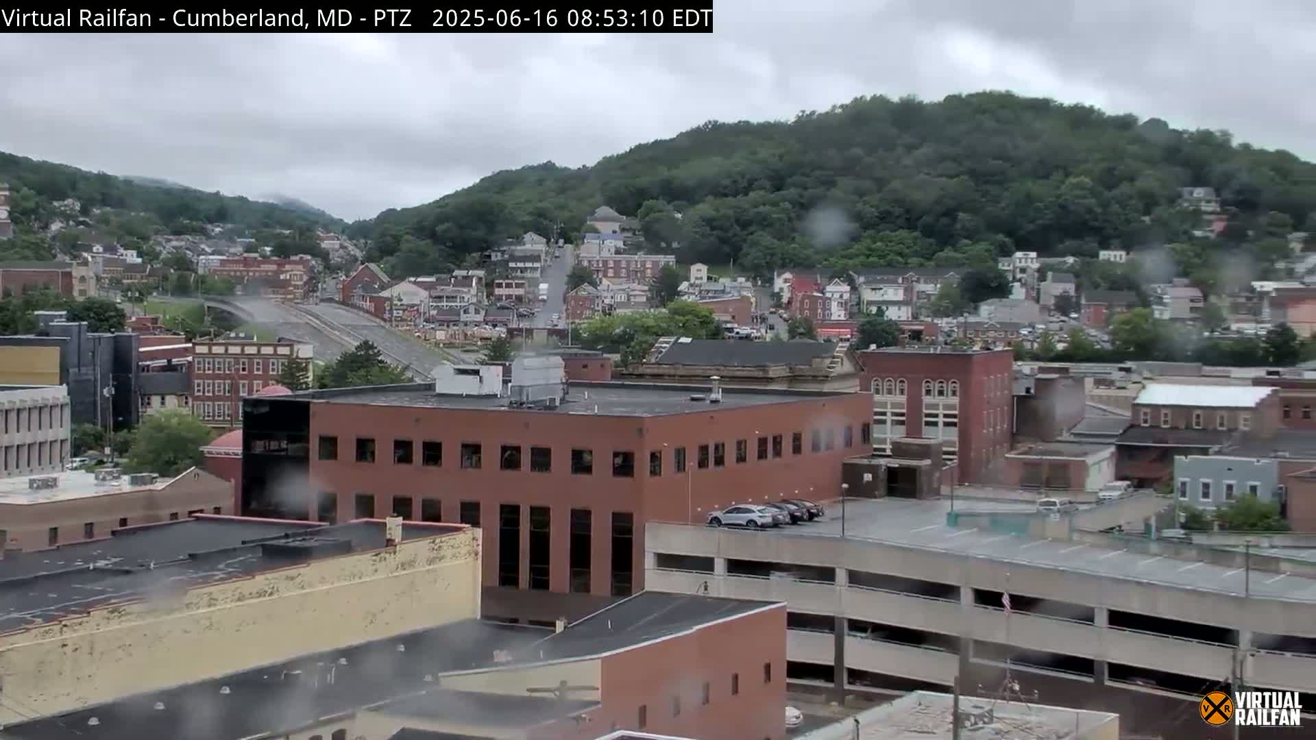 A rain-soaked view of a town nestled against a hillside, showing multi-story buildings, parking structures, and a highway overpass.