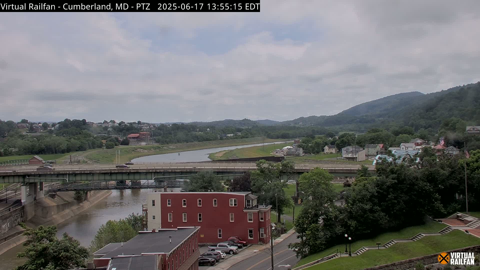 A river flows through a valley under a bridge and past a town, with overcast skies and green hills in the background.