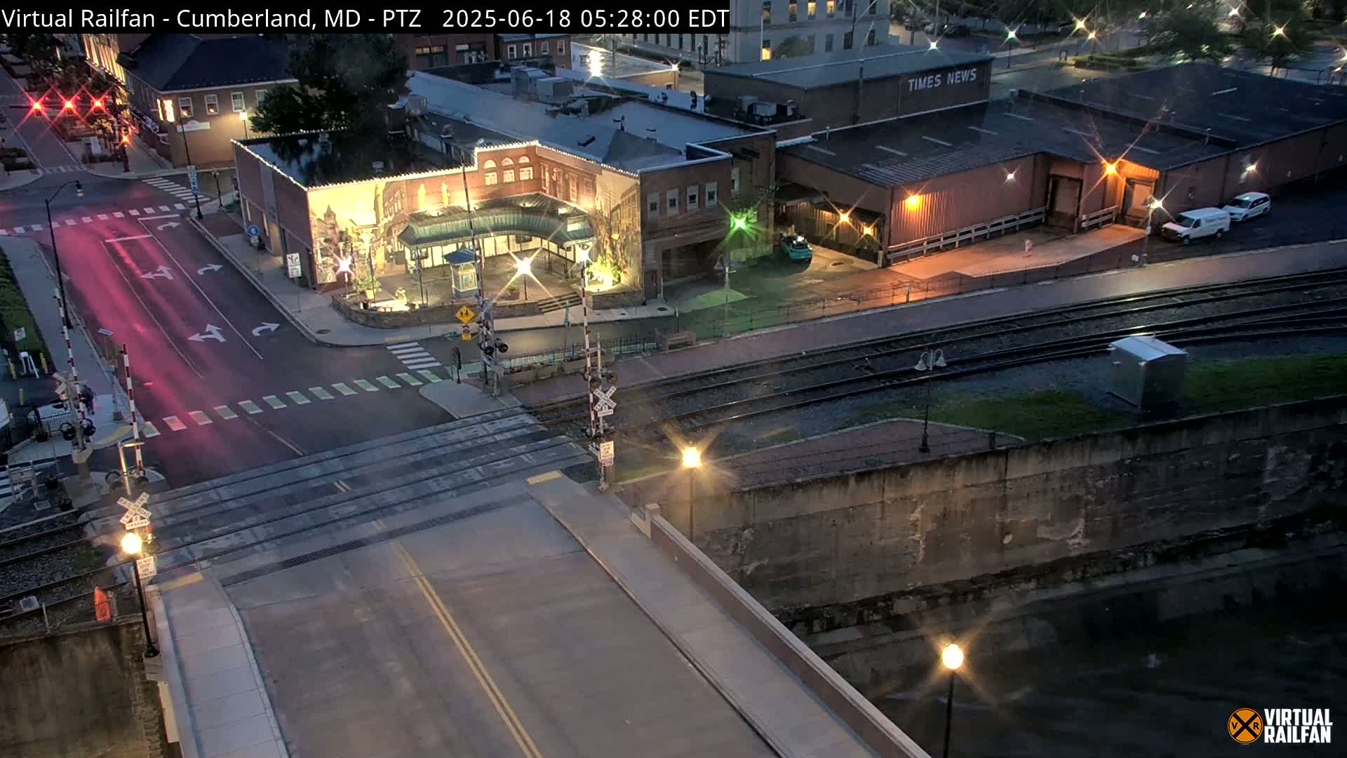 A nighttime view of a street intersection near railroad tracks, with a mural on a building and some cars parked nearby.