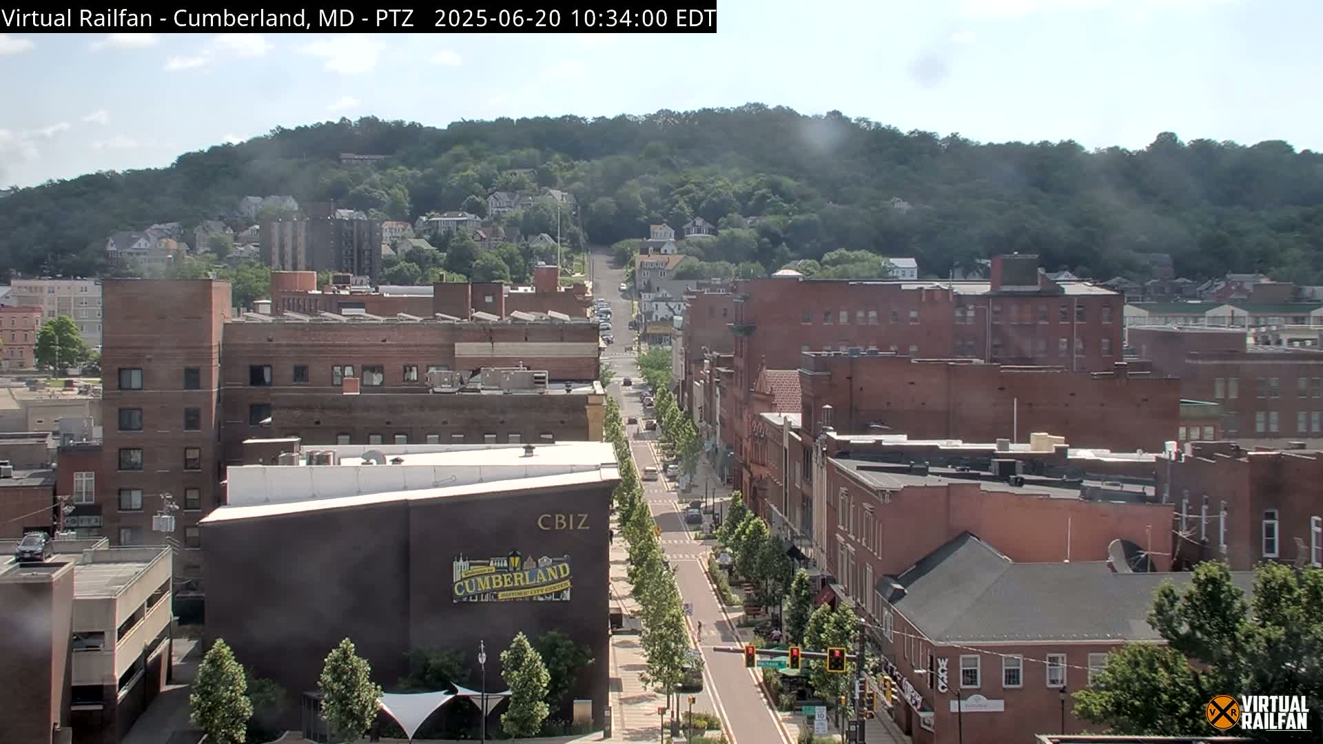 A long street lined with brick buildings and trees leads up a hill covered in green foliage on a partly sunny day.