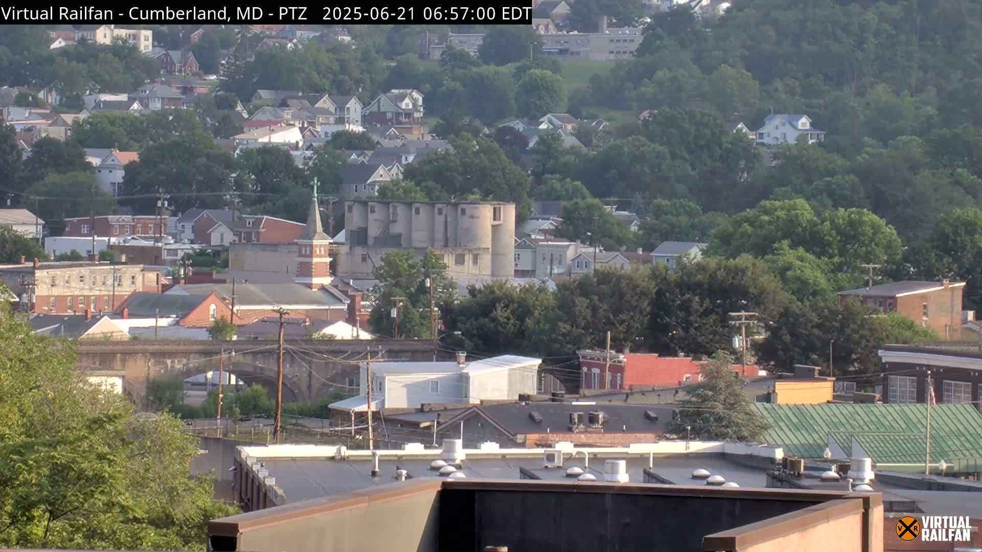 A mostly clear view of a town with several buildings, including grain silos,  set amongst green trees on a hillside.