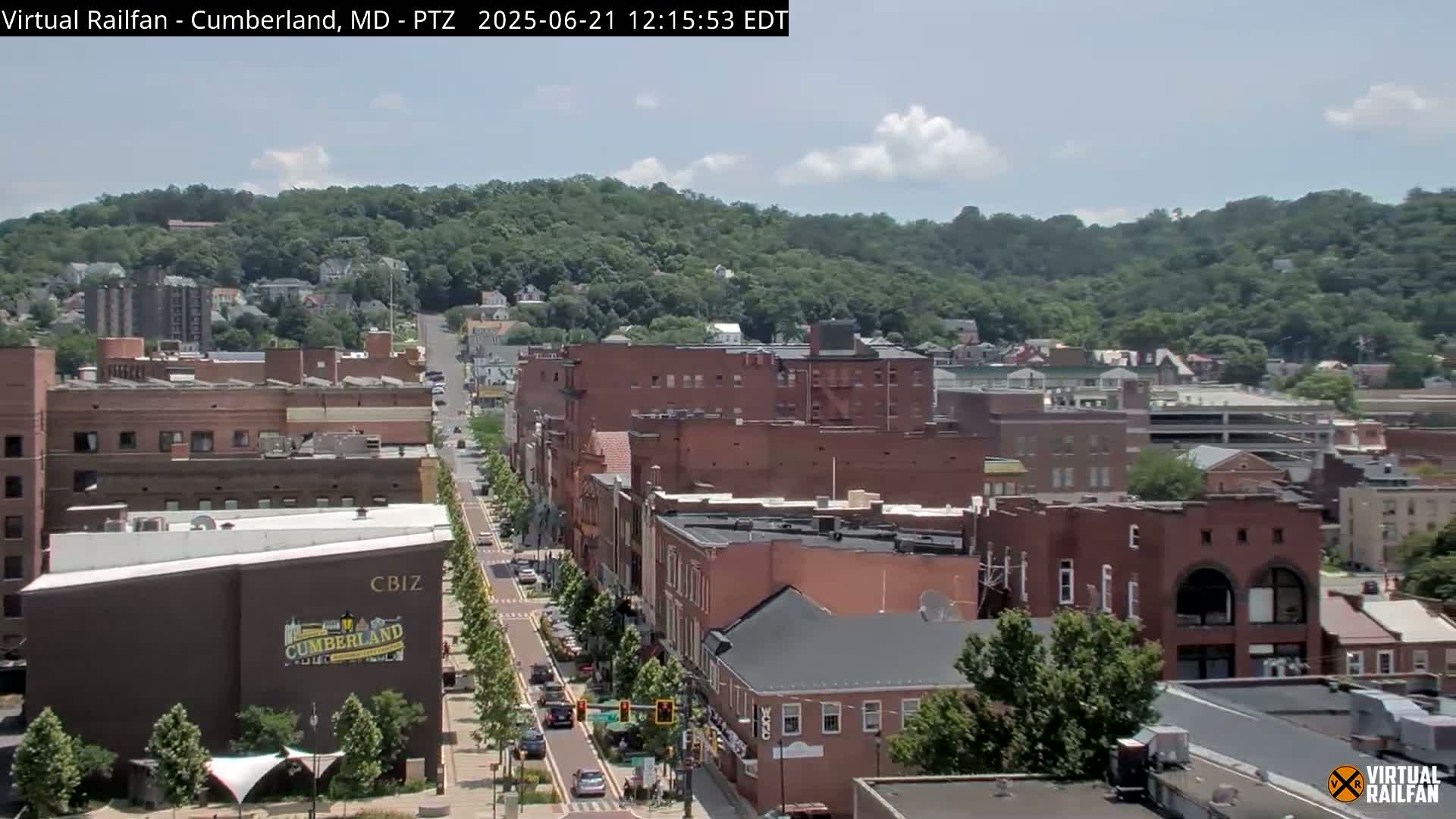 A partly cloudy summer day reveals a view of a city street lined with brick buildings, leading towards a tree-covered hill in the background.