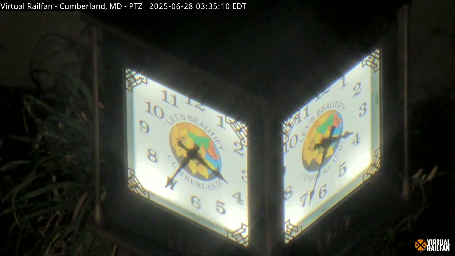 Two illuminated clocks, showing the same time, are mounted on a dark post at night next to some foliage.