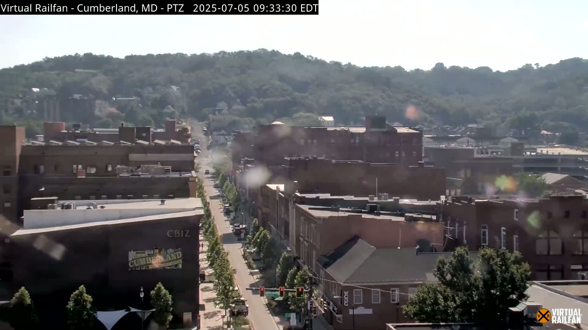 A long street lined with brick buildings stretches toward a hazy, tree-covered hill under a sunny sky.