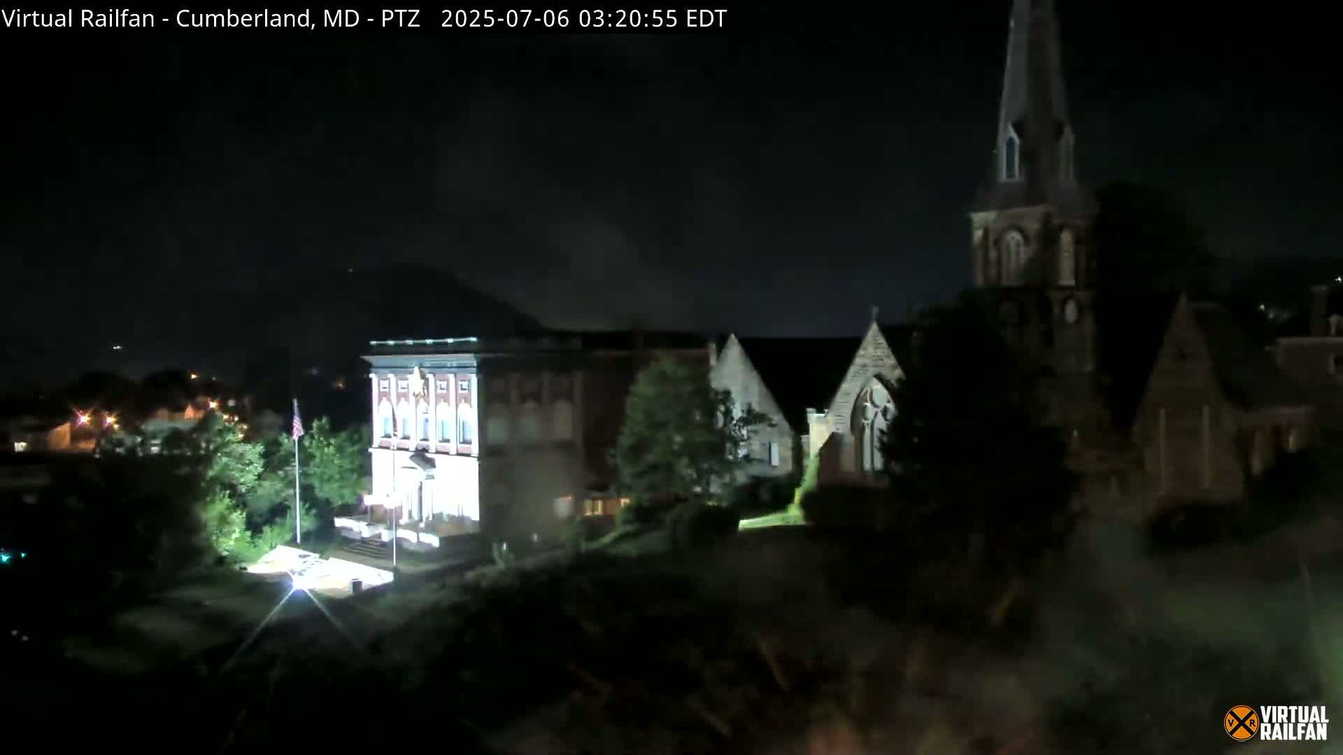 At night, a brightly lit building stands beside a church with a tall steeple, other buildings and trees are visible in the darkness.