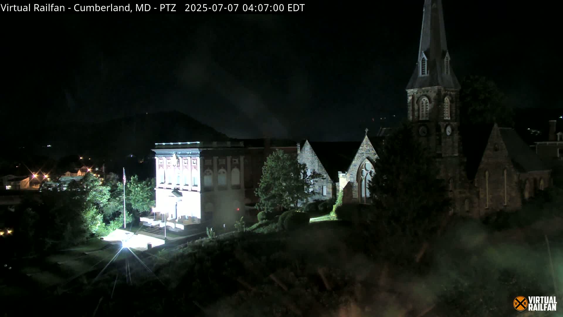 A nighttime, high-angle view shows a brick building illuminated, and adjacent stone churches, all nestled amongst trees and under a dark sky.