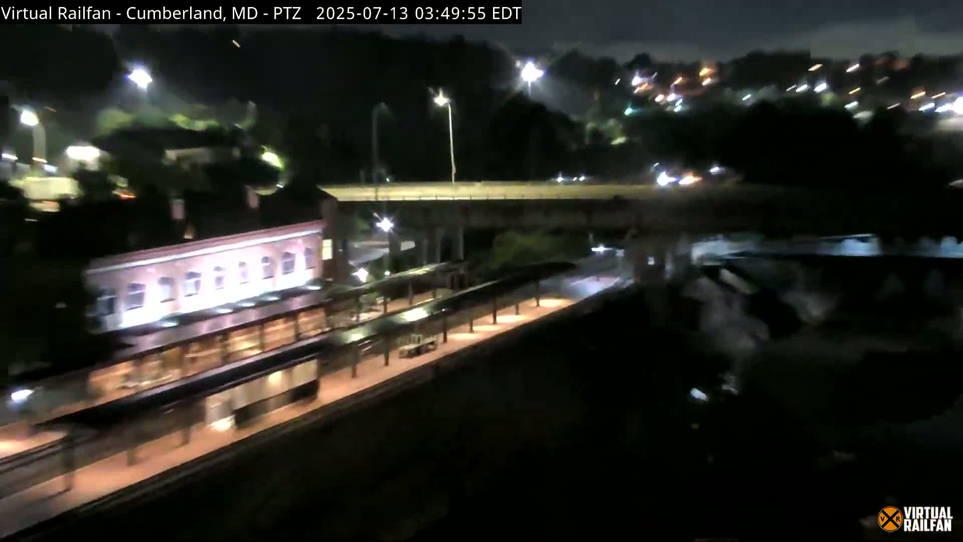 A nighttime view shows a train at a station platform next to a road with an overpass, under artificial lights.