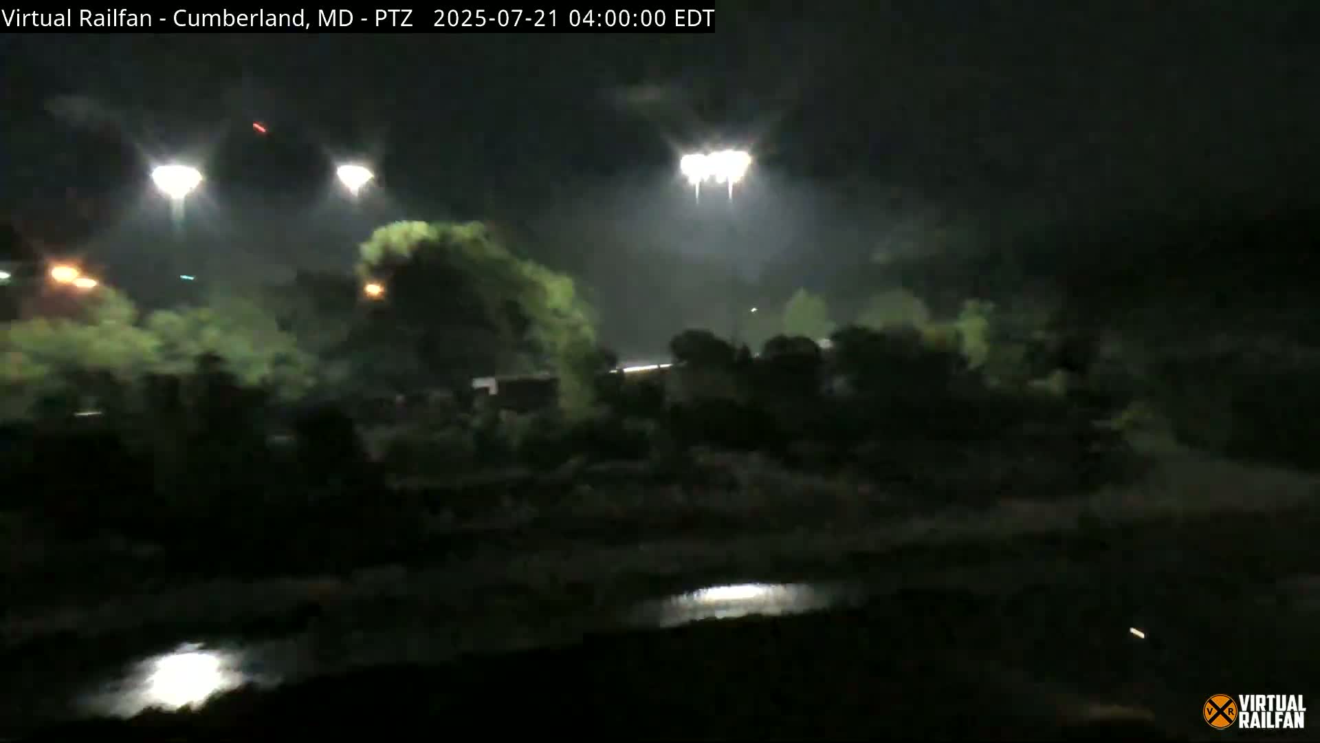 At night, under a dark sky, bright lights illuminate trees and a body of water near a roadway.