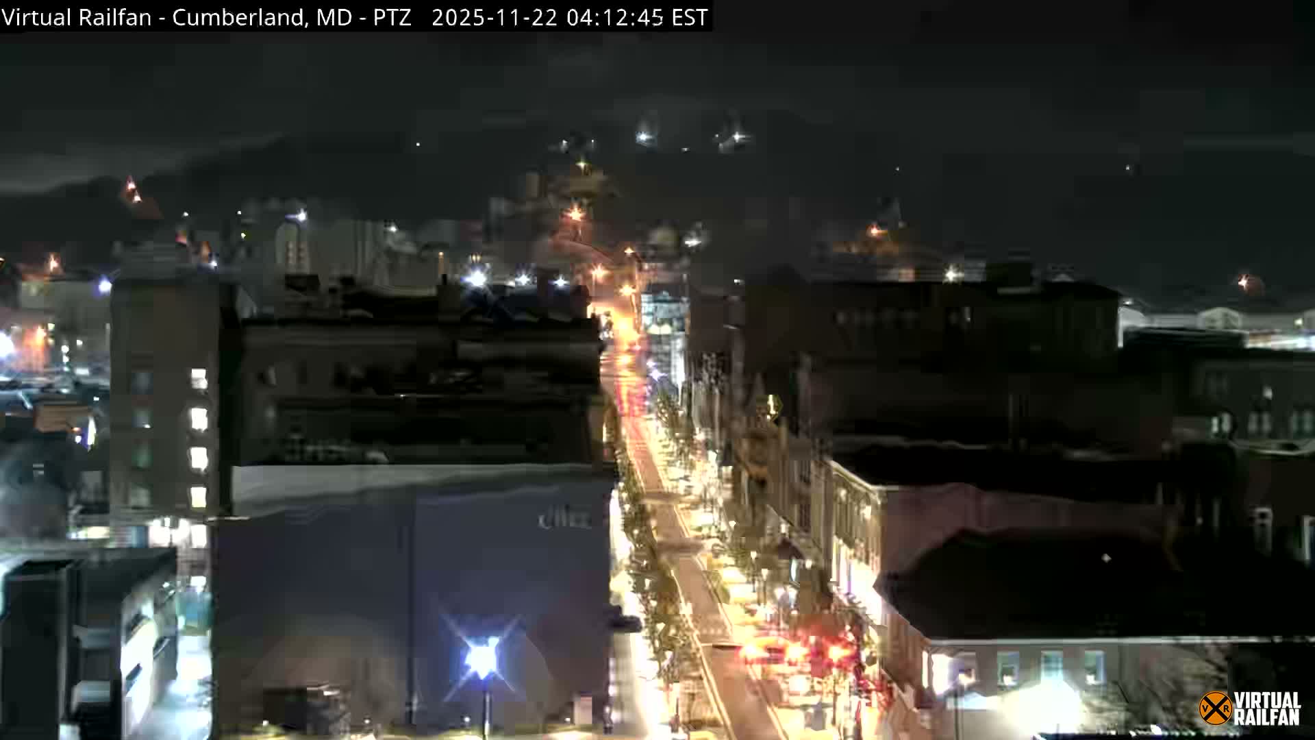 A clear night view shows a city street stretching into the distance, brightly illuminated by numerous streetlights and the lights of vehicles, flanked by multi-story buildings under a dark sky.