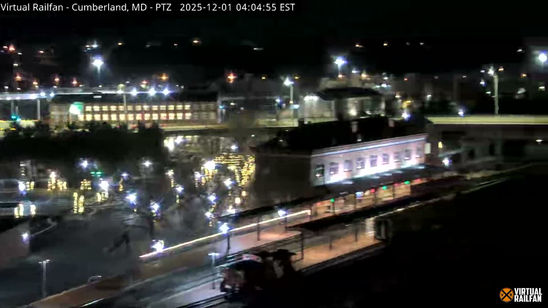 A nighttime aerial view reveals a train station with a train on the tracks, surrounded by brightly lit buildings, streetlights, and festive decorations reflected in a nearby body of water, all under clear conditions.