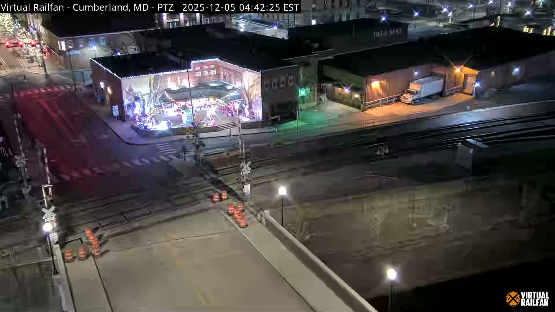 An aerial view captures a brightly lit street corner and adjacent railroad tracks in an urban area at night, featuring a building decorated with festive lights, a white semi-truck at a loading dock, and traffic cones on a clear evening.