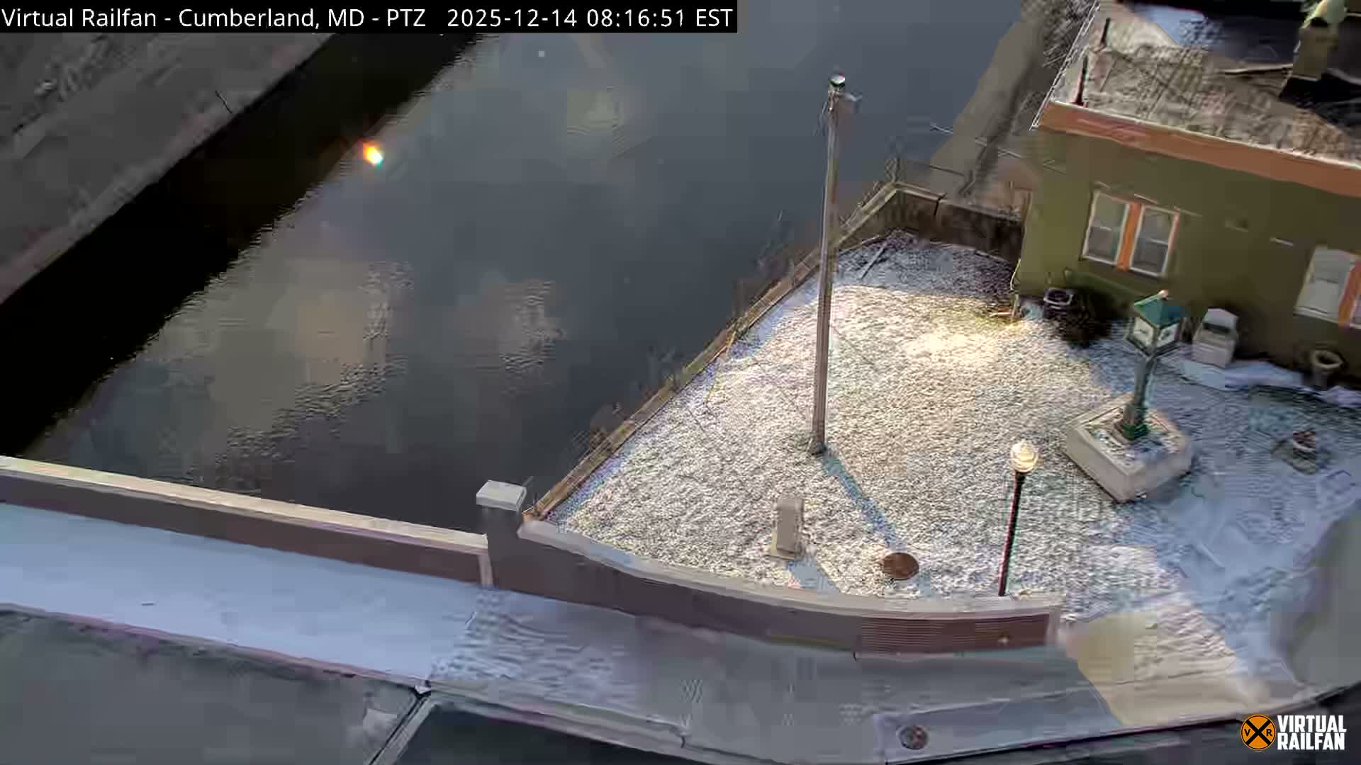 An aerial view captures a brightly lit street corner and adjacent railroad tracks in an urban area at night, featuring a building decorated with festive lights, a white semi-truck at a loading dock, and traffic cones on a clear evening.