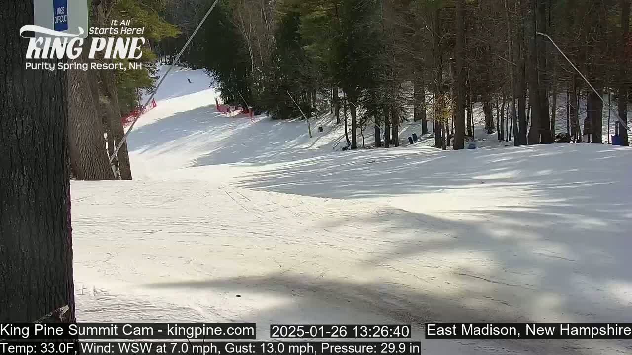A snow-covered ski slope is seen on a sunny day, with trees lining the sides.