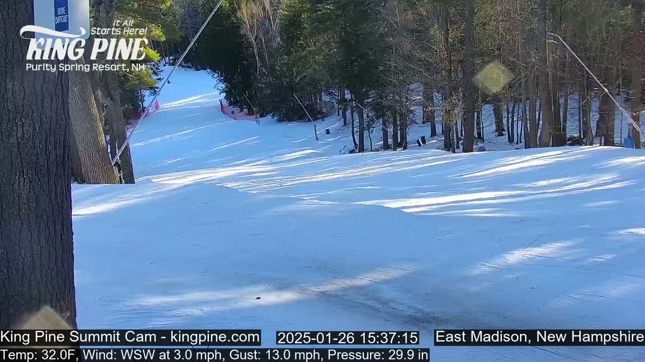 A snow-covered ski slope winds through a sunny, wooded area with a light wind.