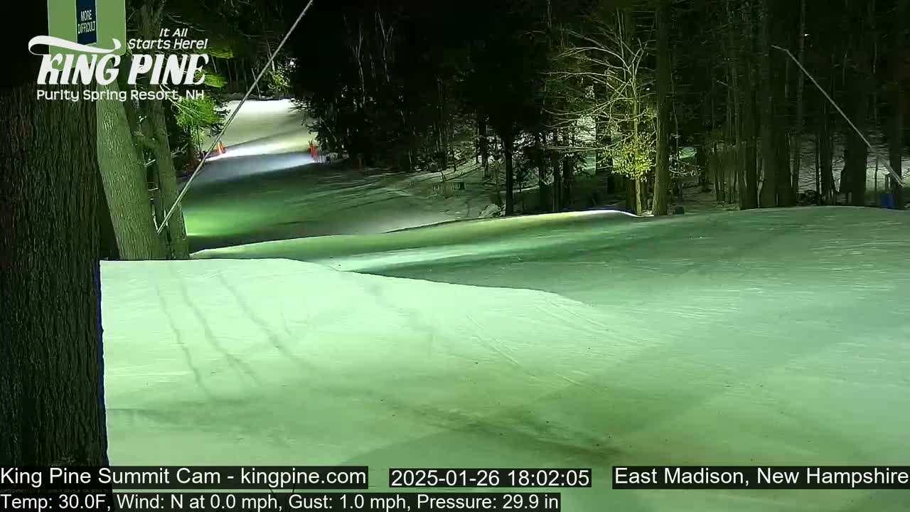 A nighttime view of a snow-covered ski slope illuminated by artificial lights, with trees lining the edges under a clear sky.