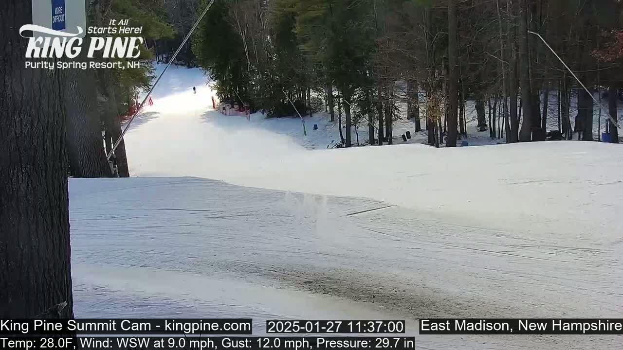 A snow-covered ski slope descends from a wooded area under a bright, sunny sky.