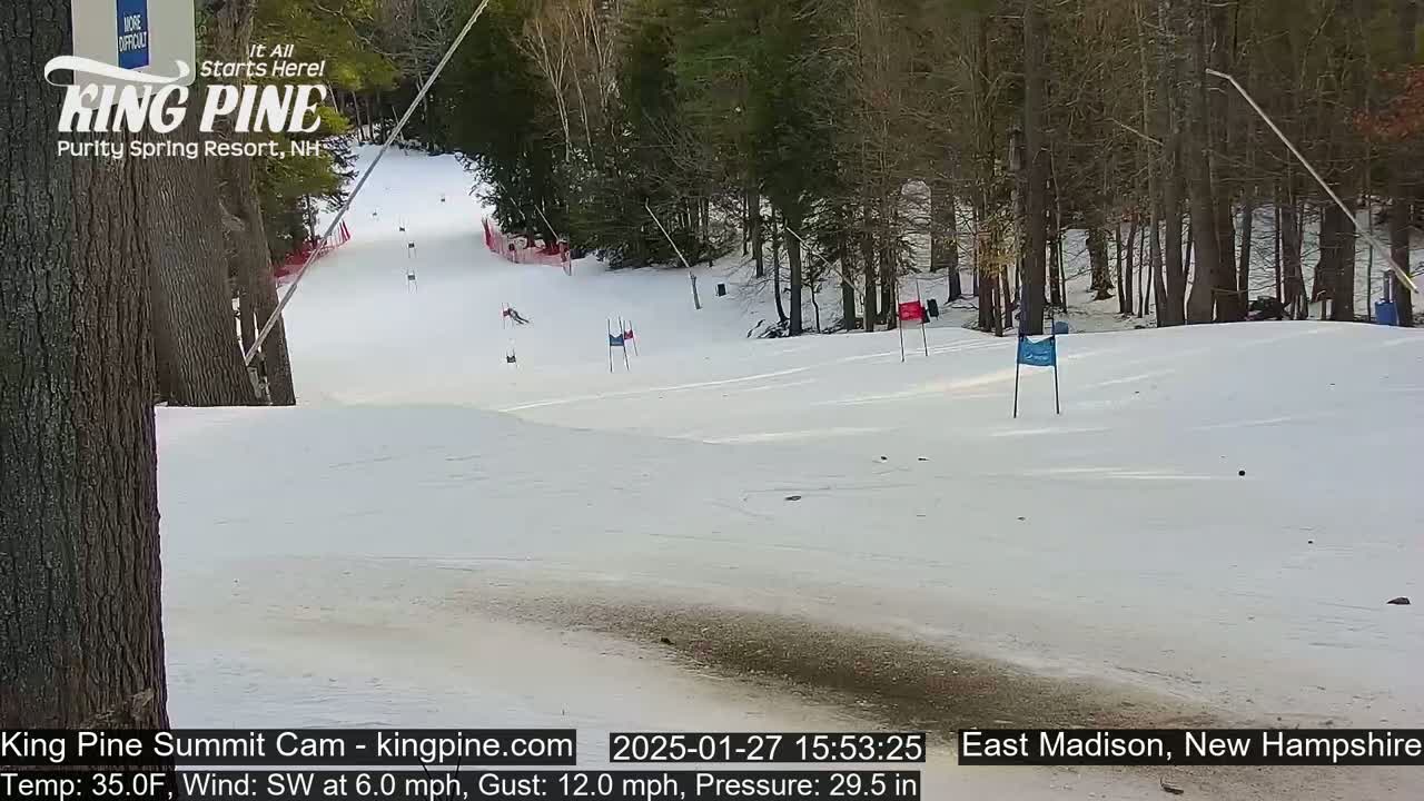 A snow-covered ski slope with several skiers visible, surrounded by trees, under a partly sunny sky with a southwest wind.