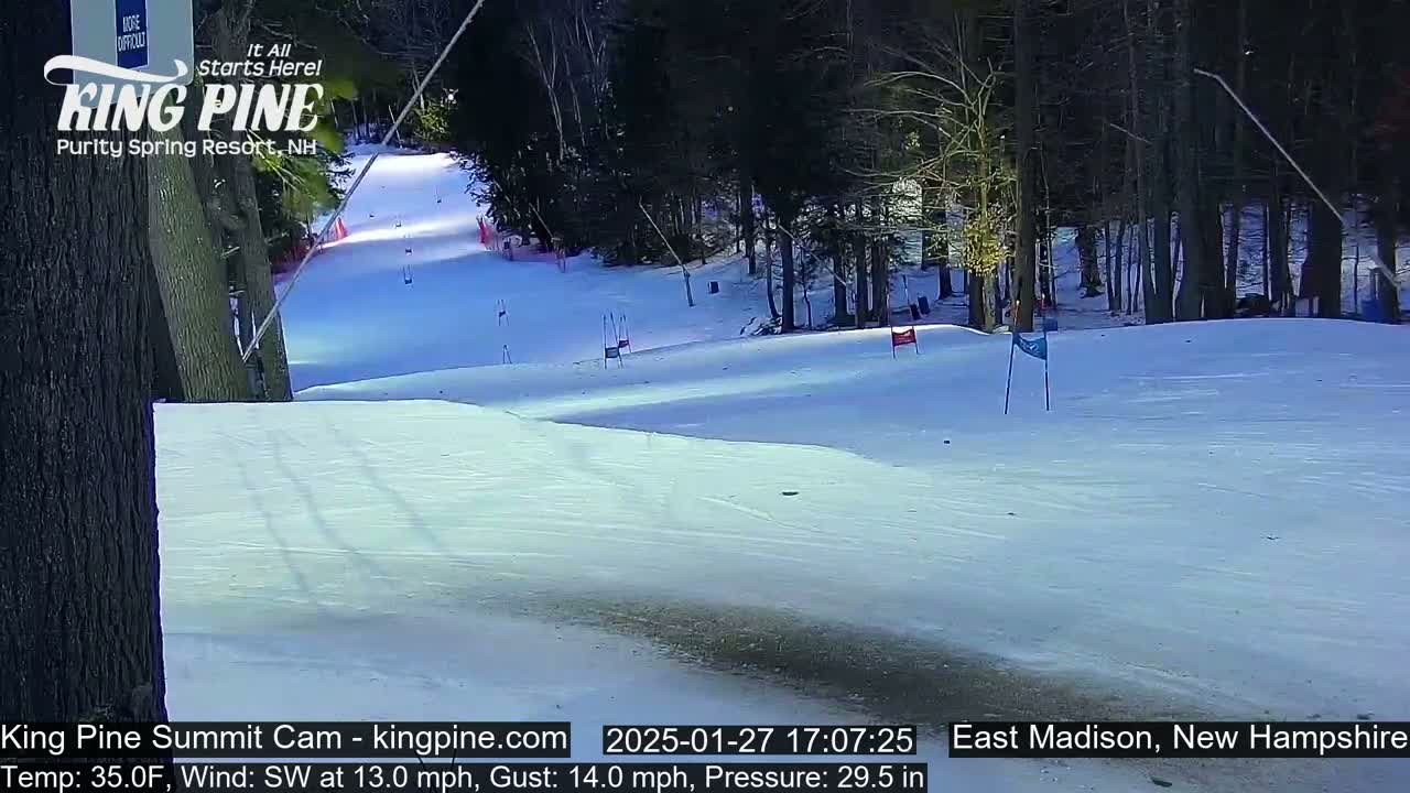 A snow-covered ski slope, partially visible through a stand of trees, is lightly used under a daytime sky with a southwest wind.