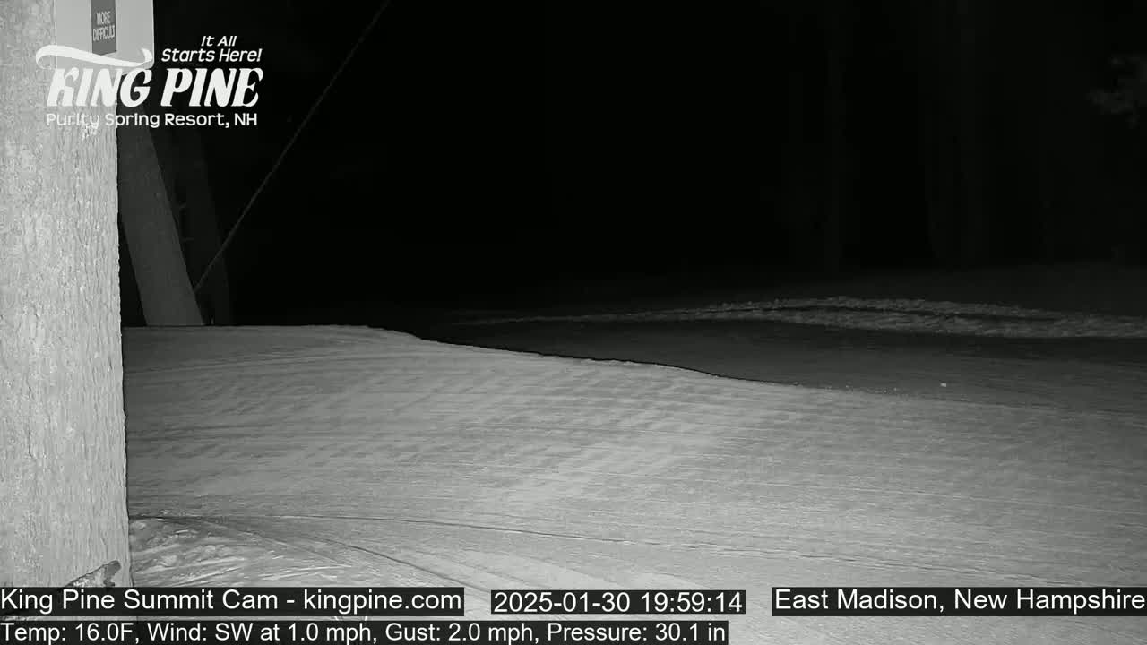 A nighttime, snow-covered scene shows a section of flat, snow-covered ground illuminated by a dim light source under a dark sky.