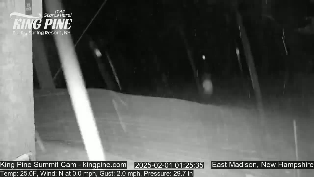 A nighttime, snowy scene shows a snow-covered ground with falling snow and a building in the background.