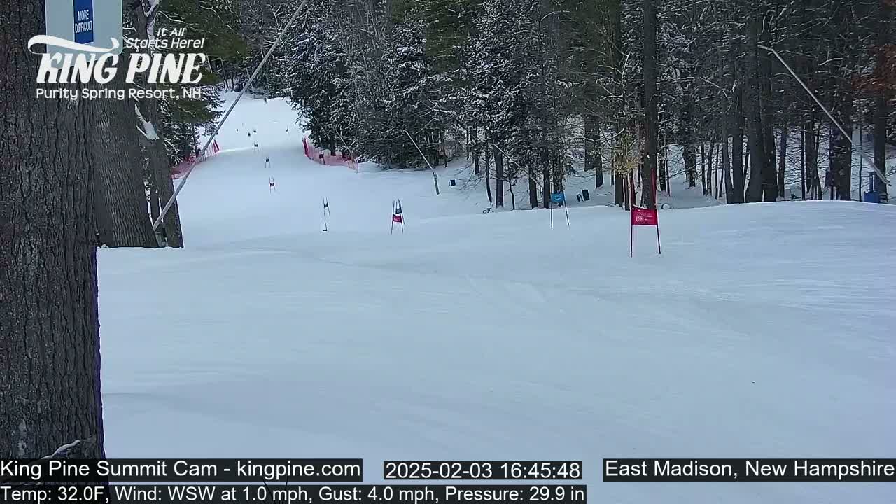 A snow-covered ski slope winds through a wooded area on a partly cloudy day.