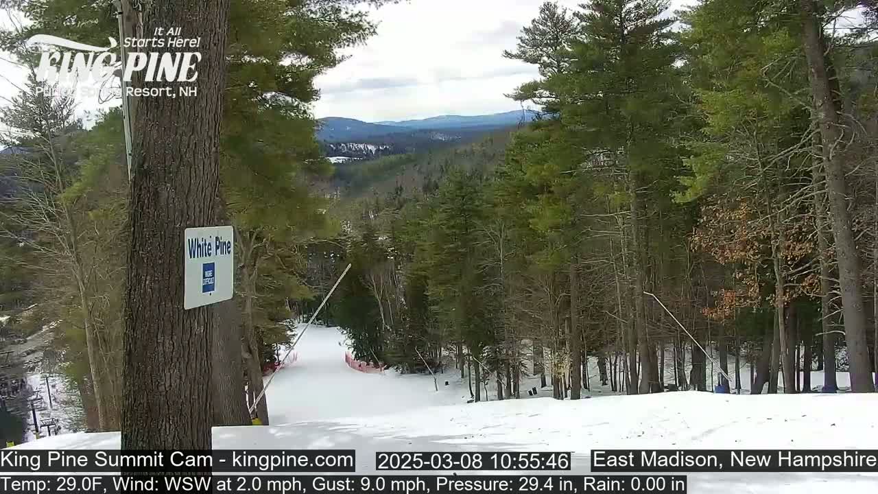A snow-covered ski slope descends through a wooded area on a partly cloudy day.