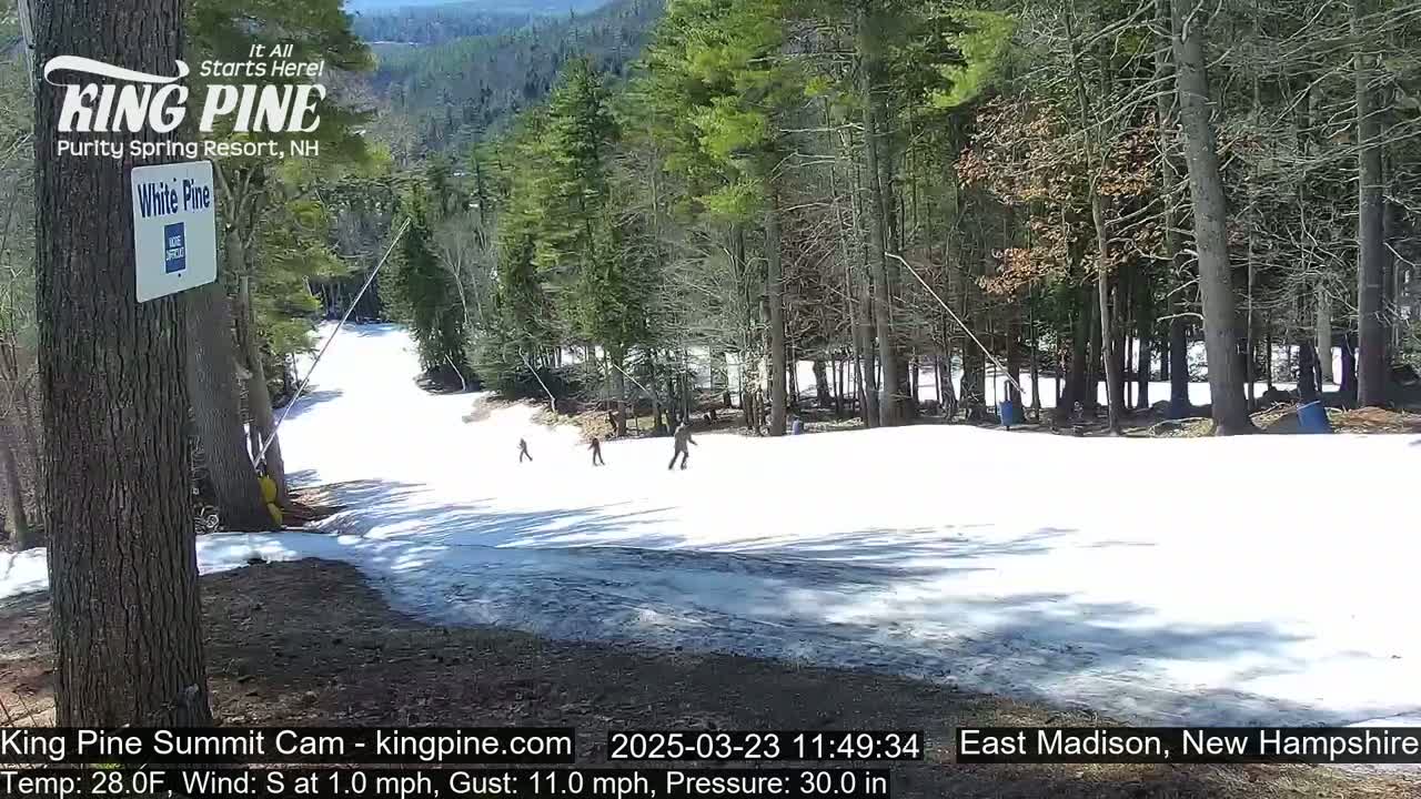 A snow-covered ski slope with a few skiers is visible amidst a partially wooded area under a partly sunny sky.