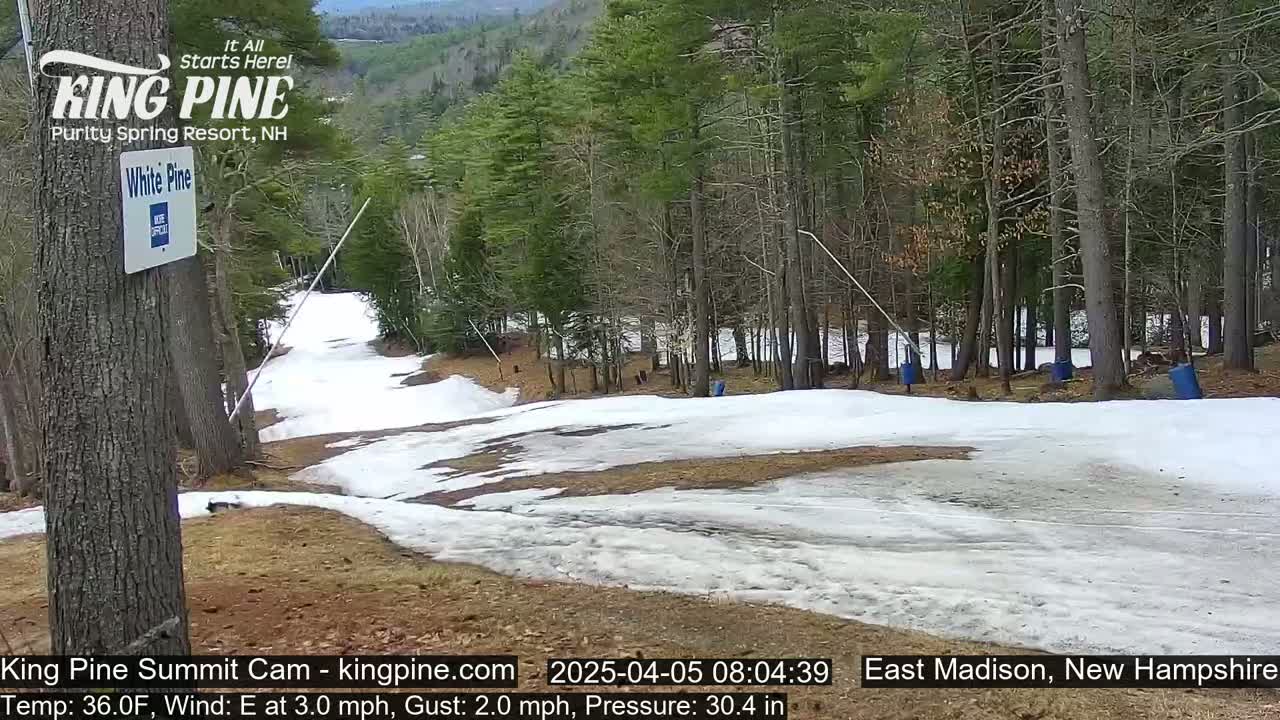 A snow-covered ski slope in a forest with patches of bare ground, under a partly cloudy sky.