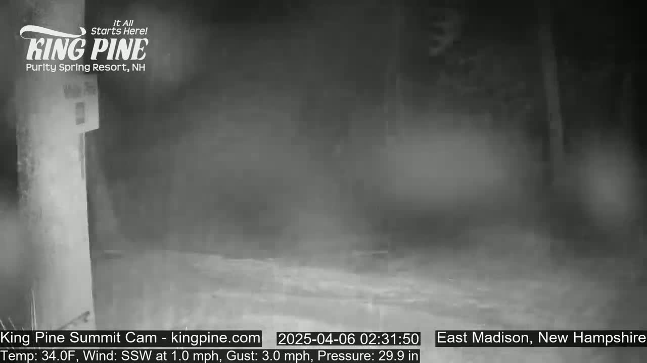 A nighttime, blurry, low-light outdoor scene shows a mostly-empty parking lot or roadway with a building in the upper-left corner, under conditions of 34°F and a 1.0 mph south-southwest wind with 3.0 mph gusts.